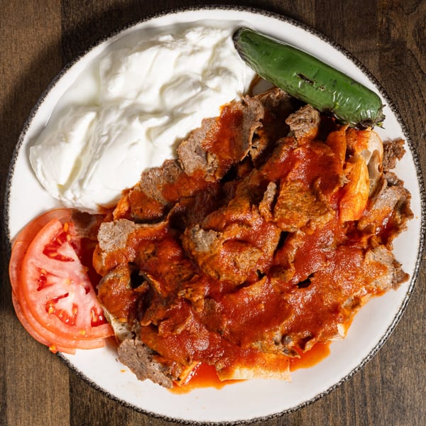 Iskender kebab with tomato sauce, yogurt, sliced tomatoes, and a green pepper on a white plate.
