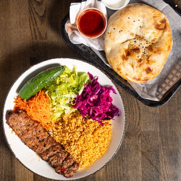 Turkish Adana kebab plate with bulgur, salad, grilled pepper, and a side of pita bread and sauces.