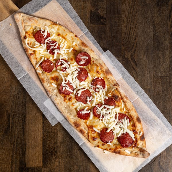 A Turkish pide, boat-shaped flatbread, topped with pepperoni and shredded mozzarella cheese on parchment paper.