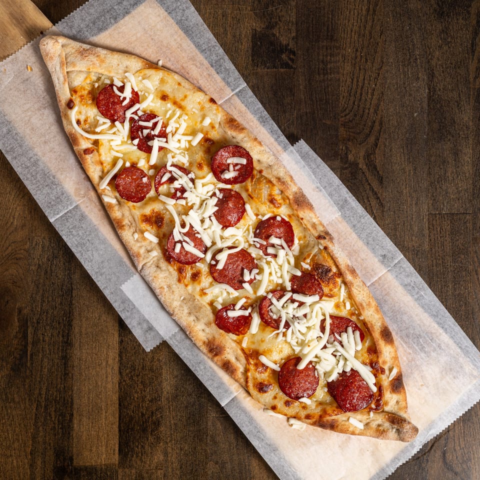 A Turkish pide, boat-shaped flatbread, topped with pepperoni and shredded mozzarella cheese on parchment paper.